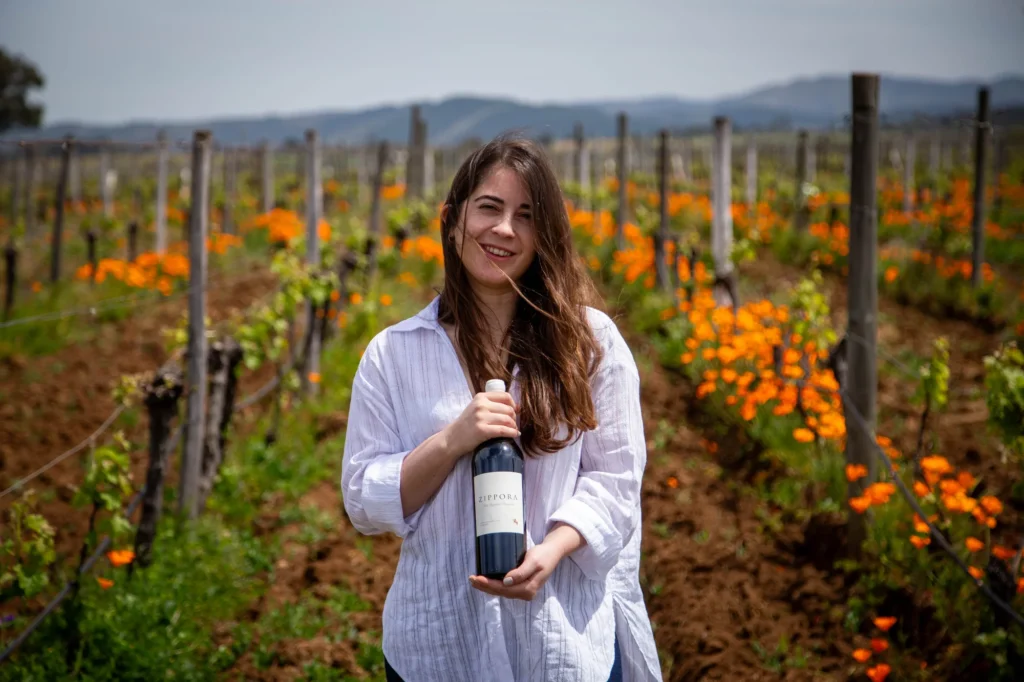 La imagen demuestra el giro de timón experimentado en la bodega Viña Odfjell, mostrando a su enóloga en una viña ecológica llena de flores con el nuevo vino de El Maule entre sus manos.